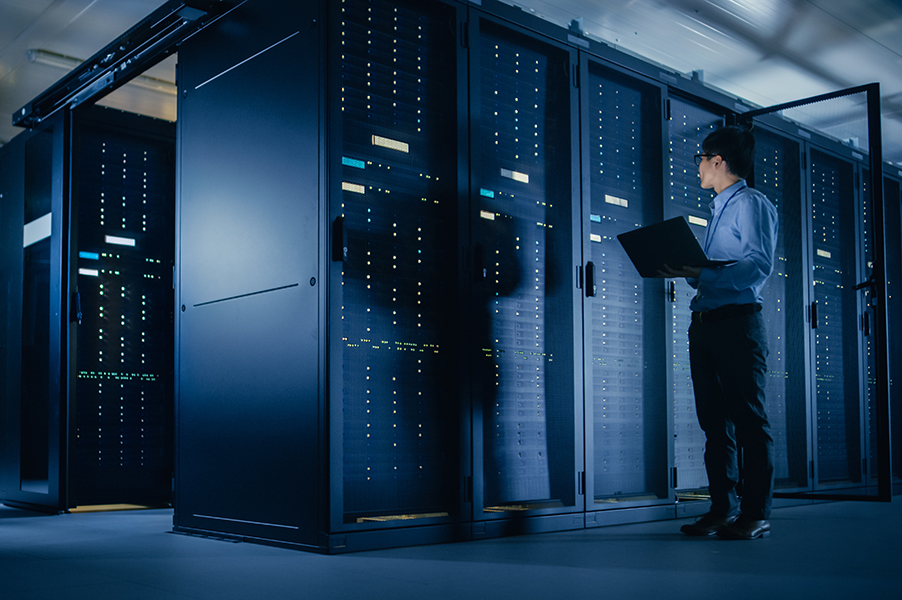 In Data Center: Male IT Technician Running Maintenance Programme on a Laptop, Controls Operational Server Rack Optimal Functioning.